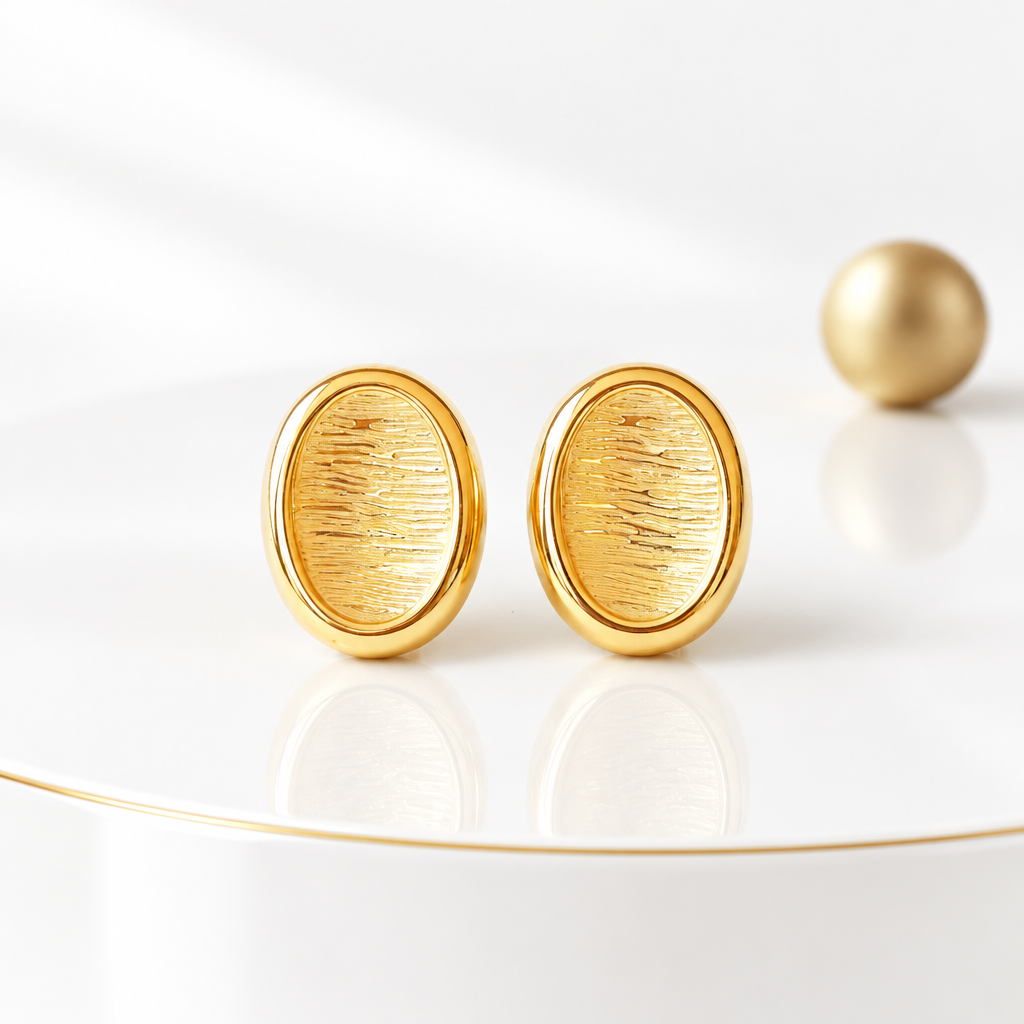 Textured oval button stud earrings in 18k PVD Gold on a glossy white podium with a matte gold sphere in the background